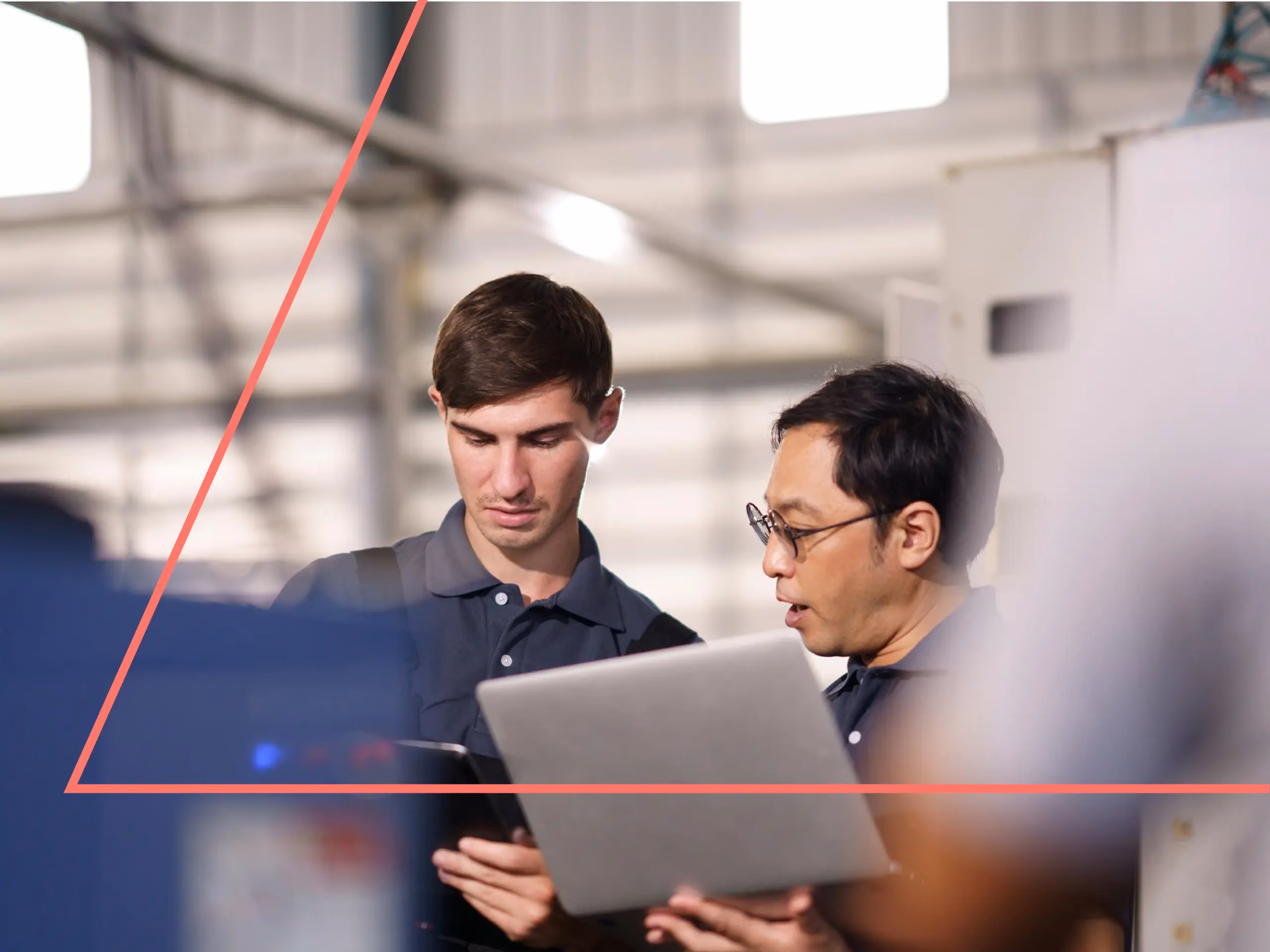 Two men in an industrial facility look at a laptop, one holding the device while the other speaks.
