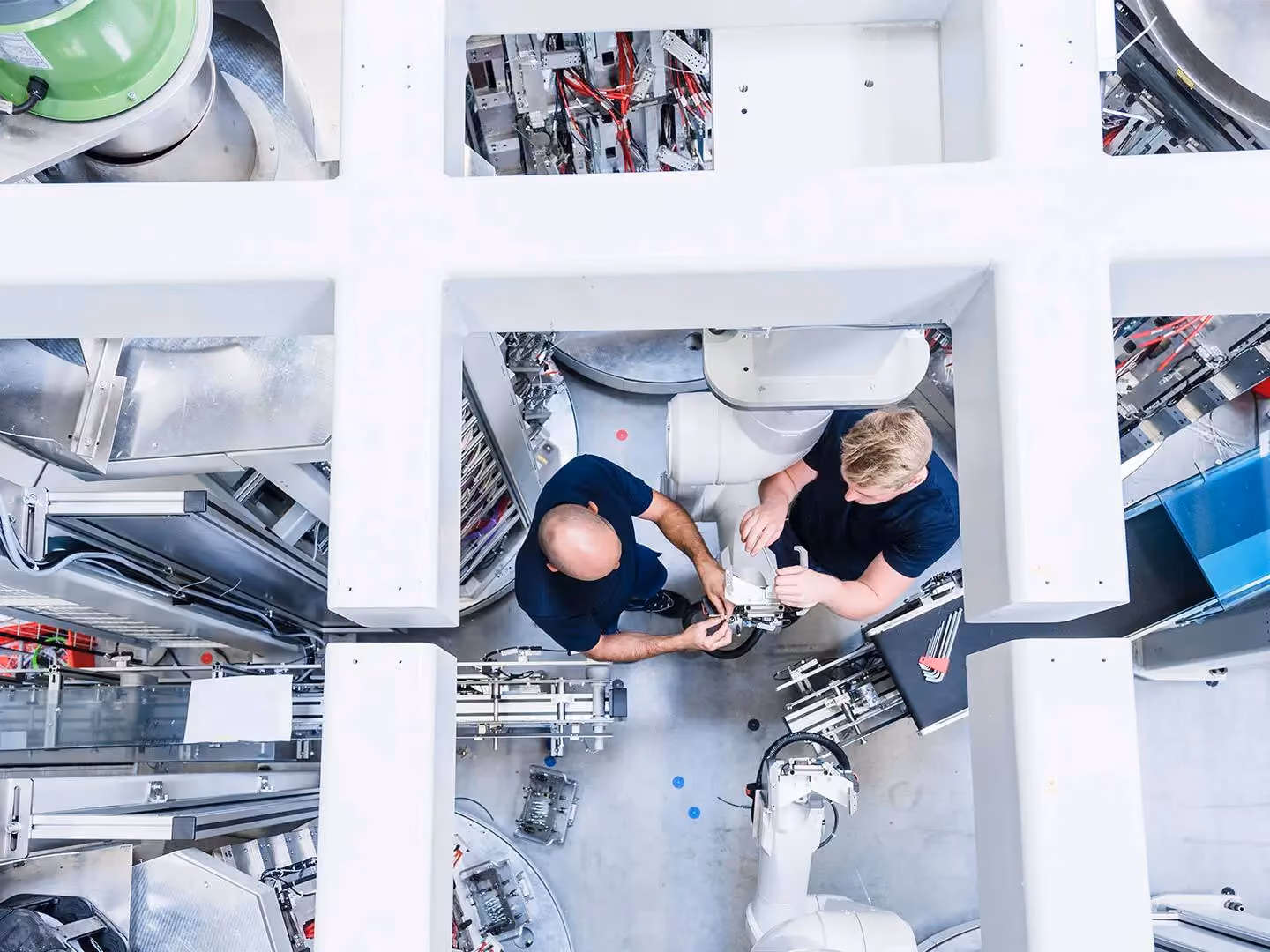 Overhead view of two technicians working on a robotic arm in a factory with white structural framework.