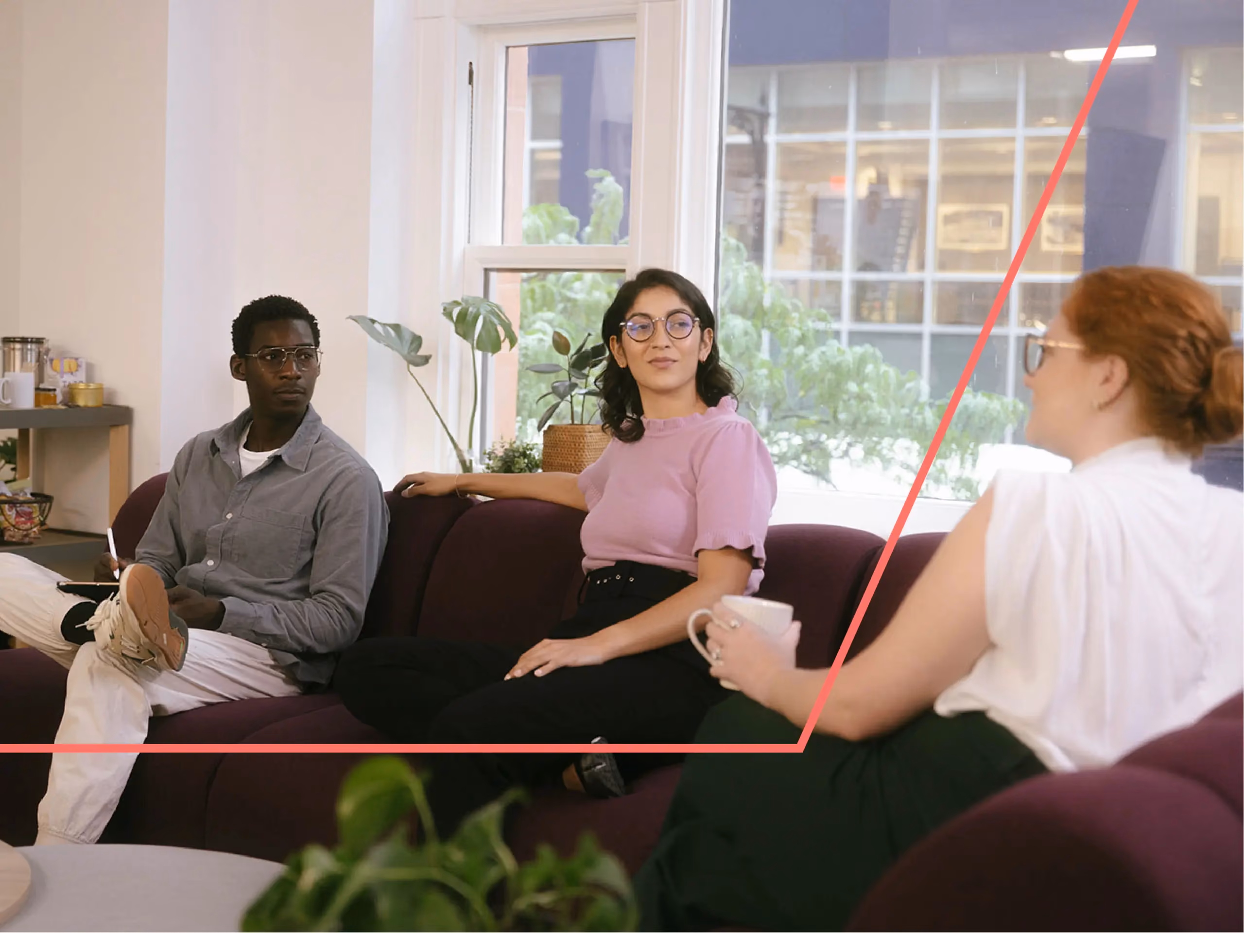 Three people sitting on a maroon couch in an office lounge, talking, near a window with plants.
