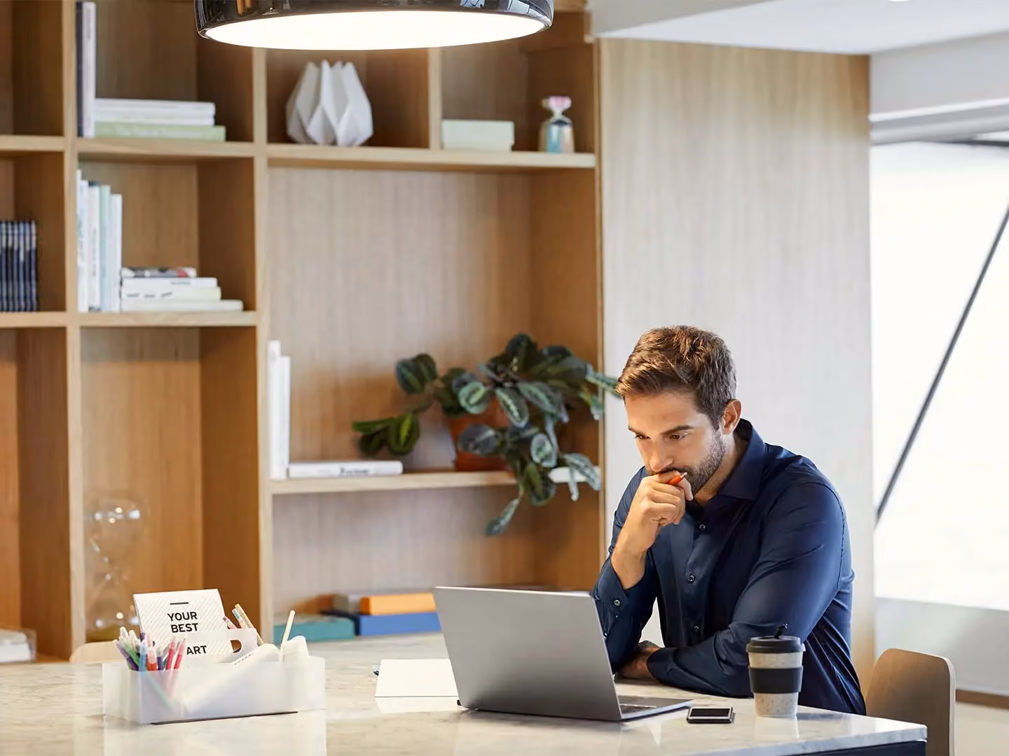Man in navy shirt sits at desk with laptop, hand on chin, in office with bookshelves and a coffee cup nearby.