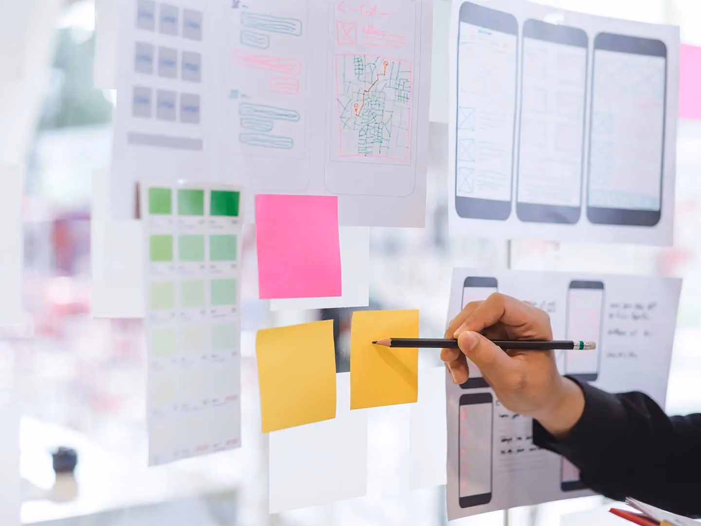 Hand holding a pencil near sticky notes and app wireframe sketches pinned to a glass wall.
