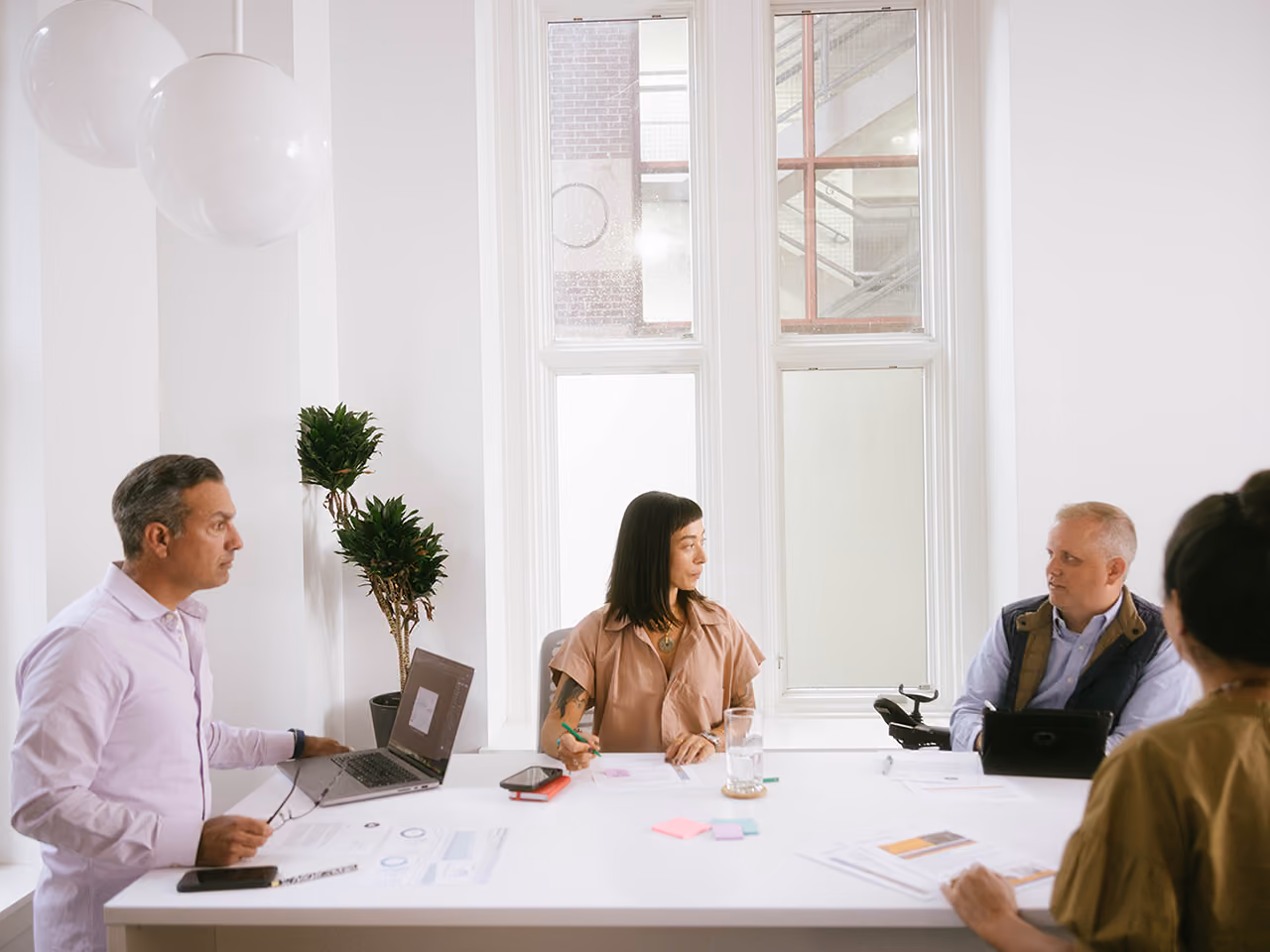 Four colleagues sit at a white conference table with laptops, papers, and sticky notes during a meeting near large windows.