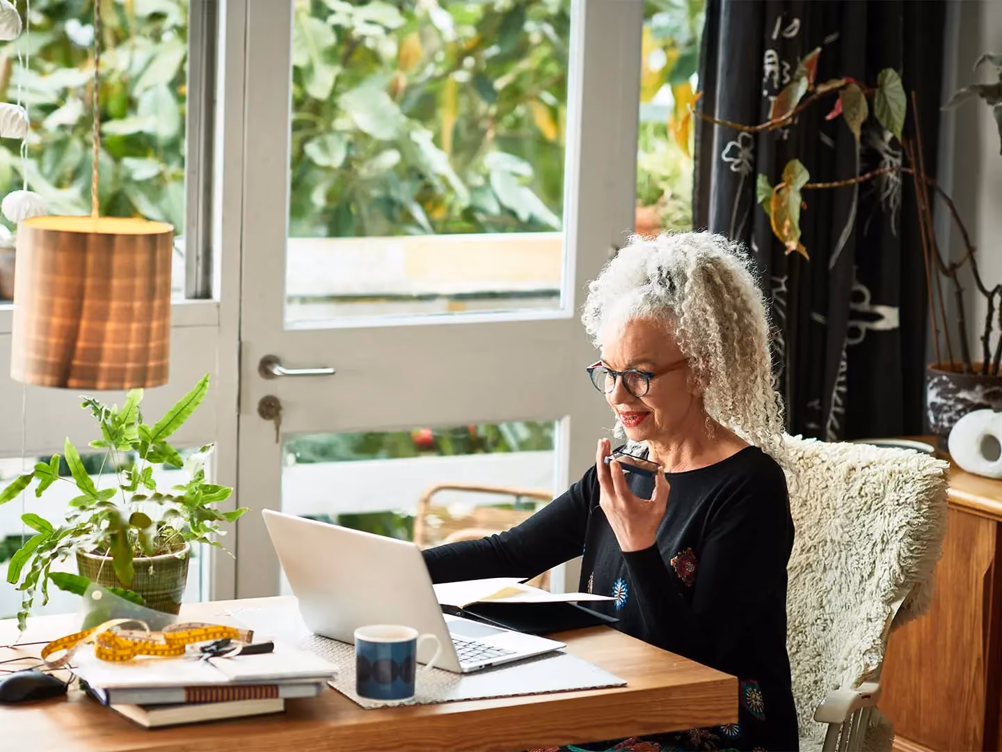 Woman with curly gray hair sits at a desk using a laptop while speaking into a smartphone.