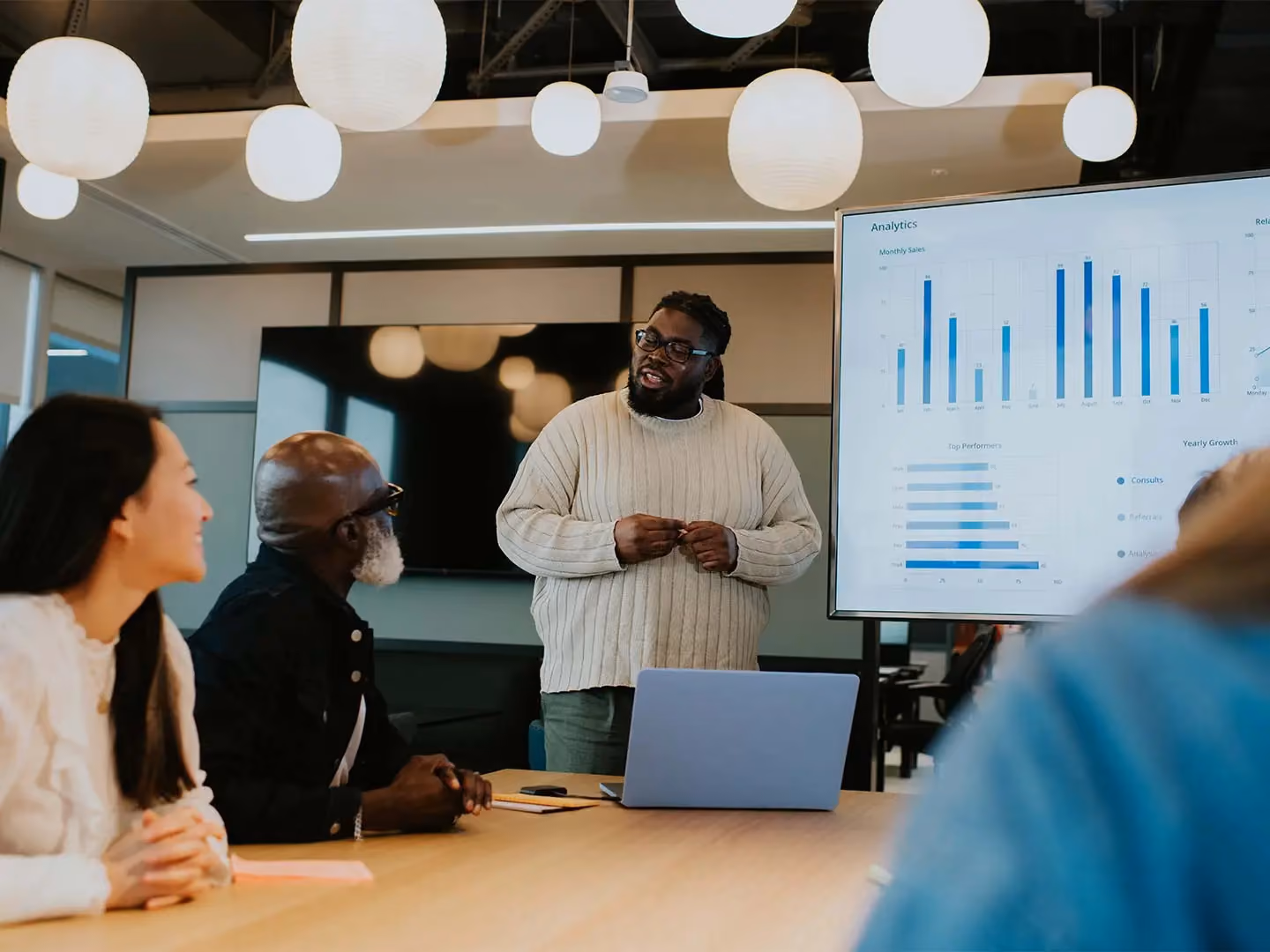 Man presenting analytics dashboard with sales charts on screen to colleagues seated at conference table.