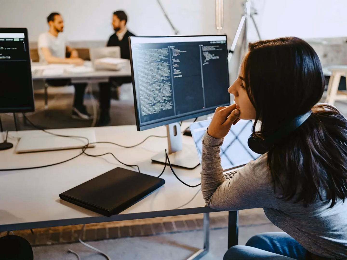 Woman with headphones around her neck views code on a monitor at a desk, with two colleagues talking in the background.