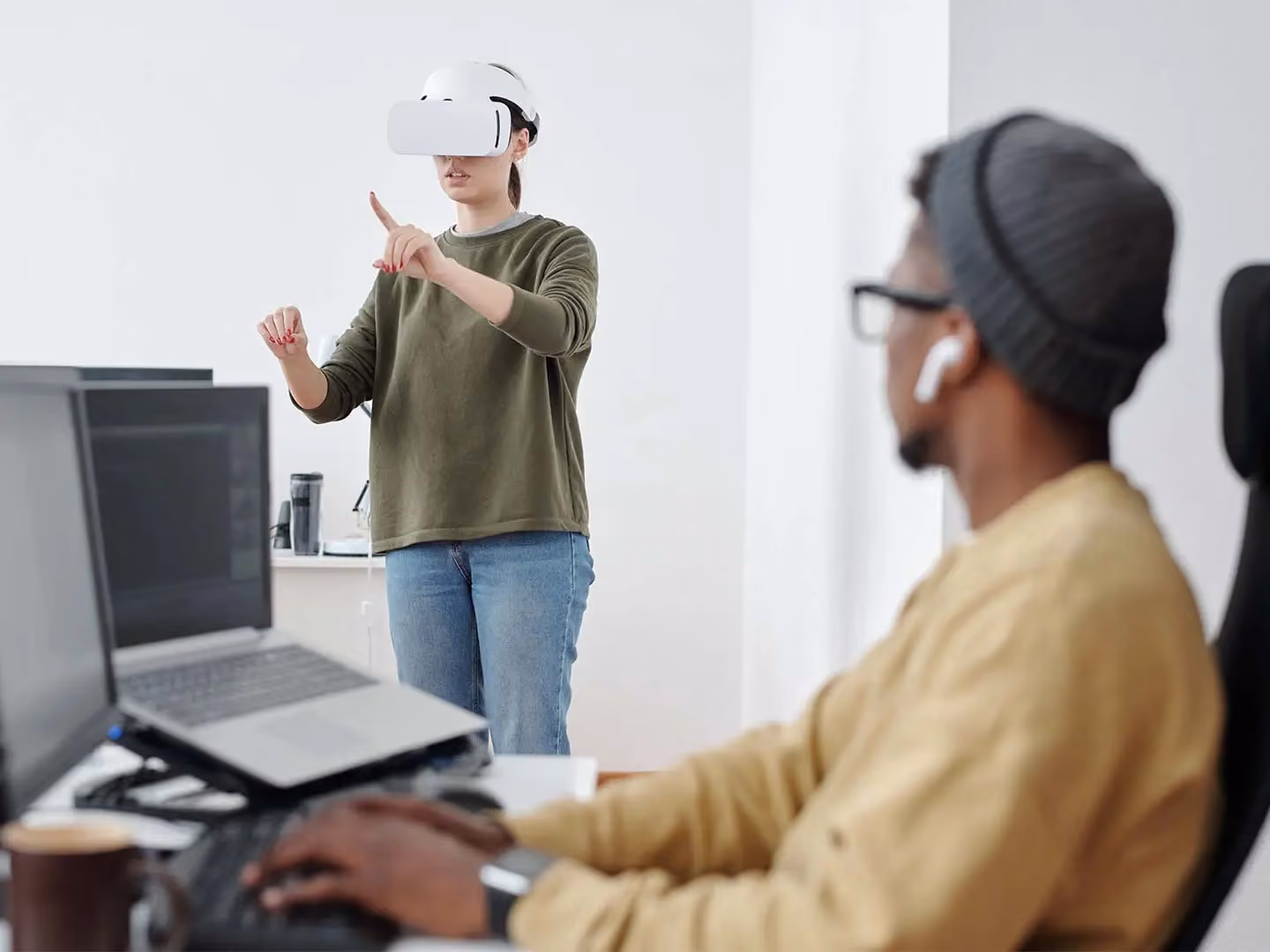Woman wearing a VR headset gestures with her hands while a man in the foreground works on a laptop.