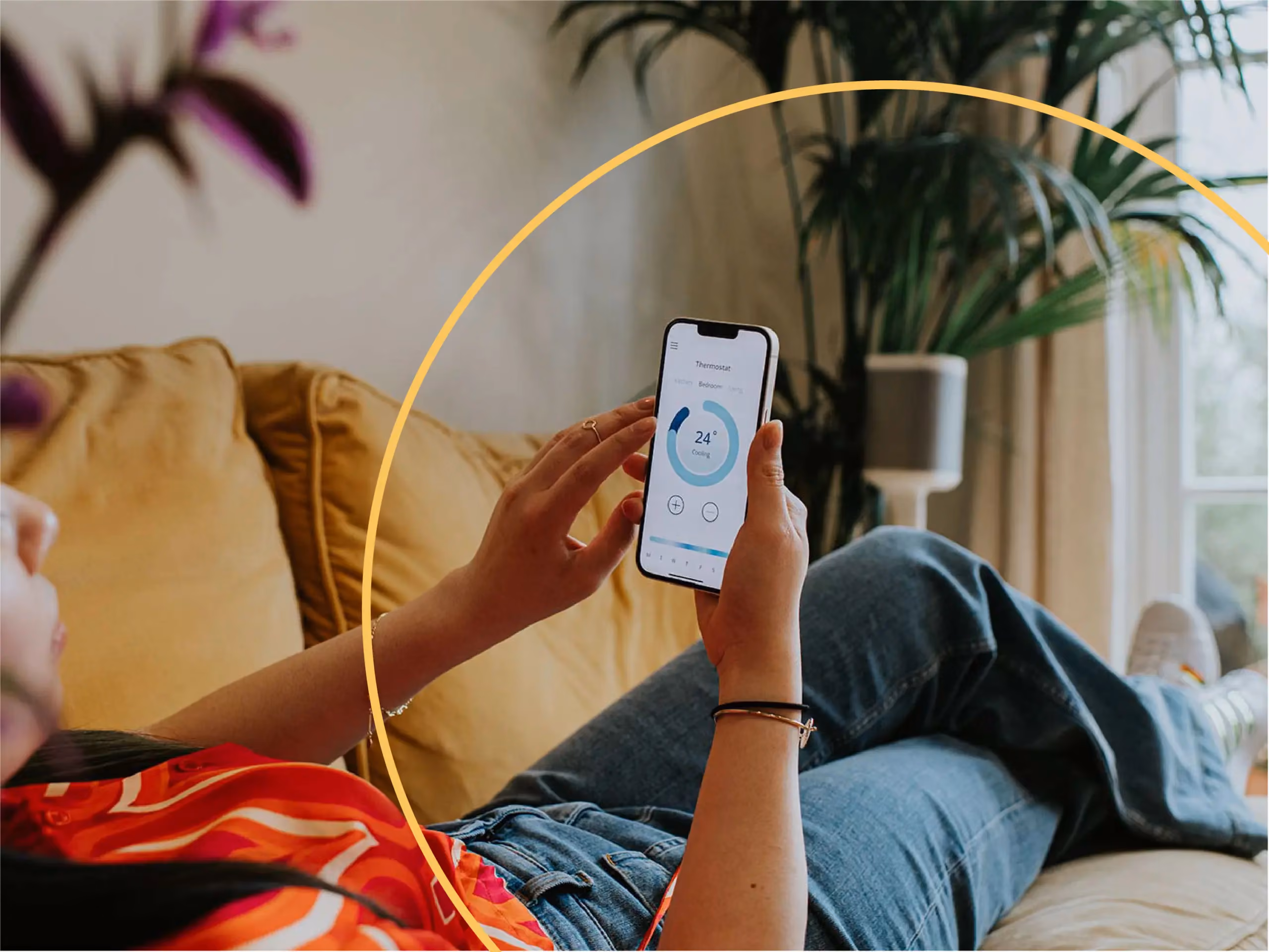 Person reclining on a couch adjusts a smart thermostat app on a smartphone, showing 24 degrees cooling.
