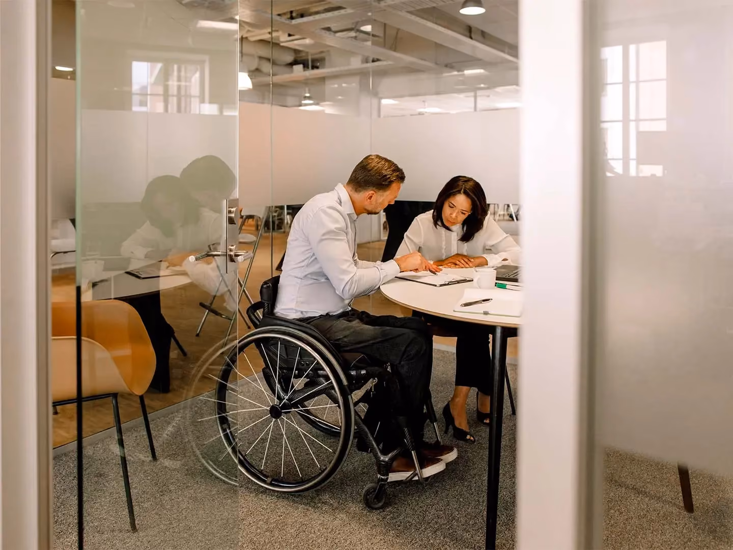 Man using a wheelchair and a woman review documents together at a round table in a glass-walled office.