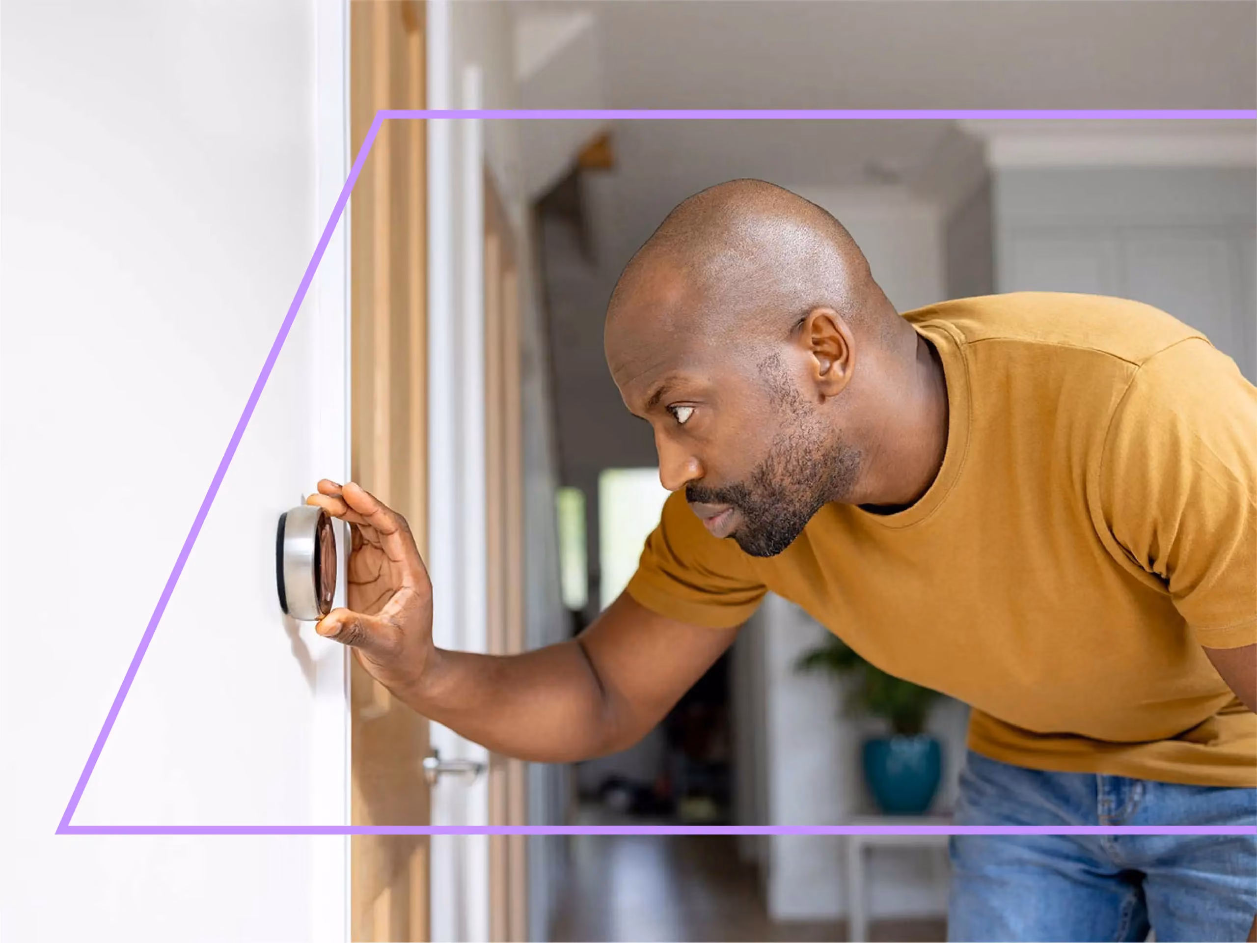 Man in a mustard shirt leans in closely to adjust a round smart thermostat on a wall.