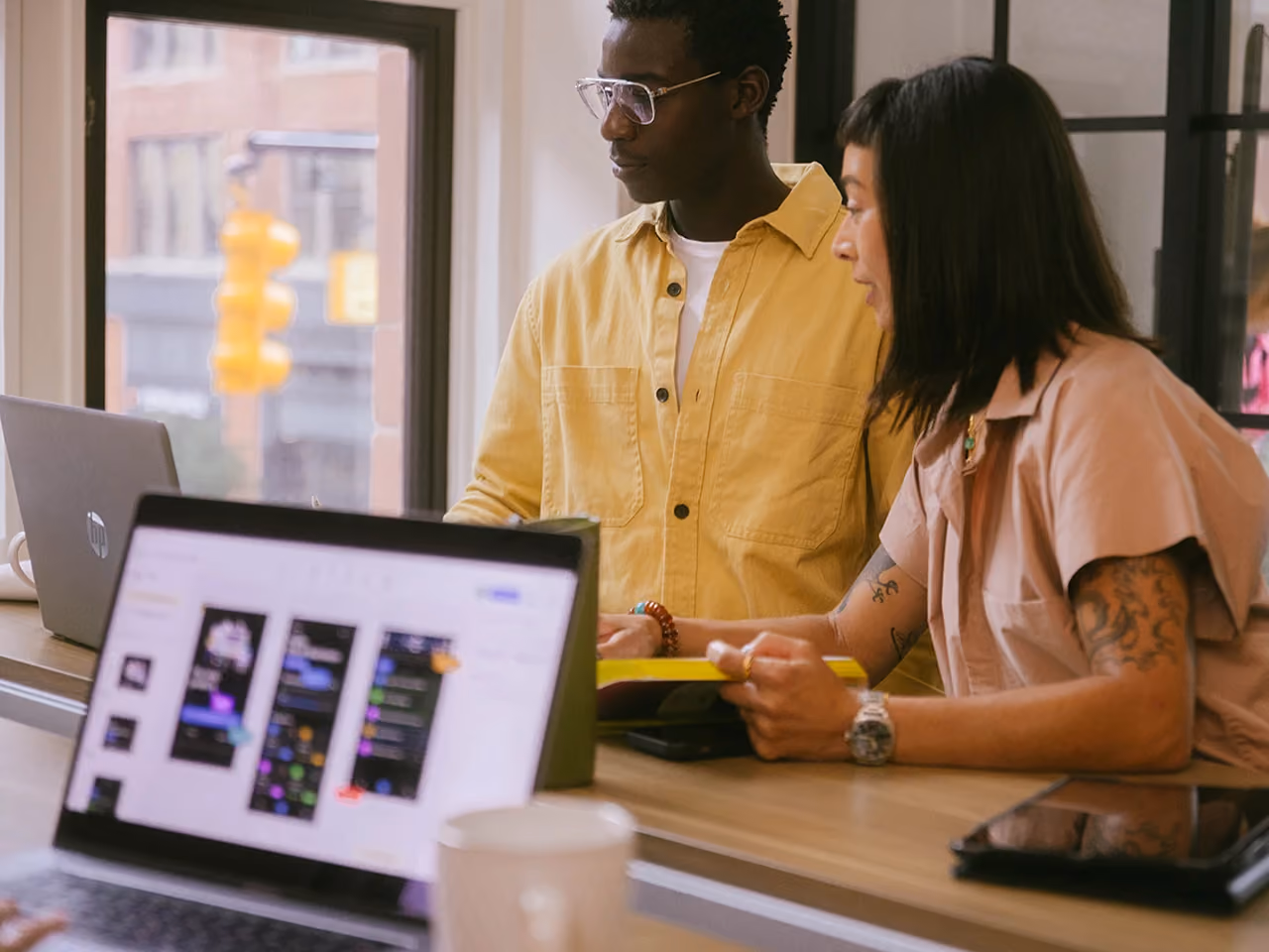 Two colleagues review a laptop screen together at a wooden desk in an office with a large window.