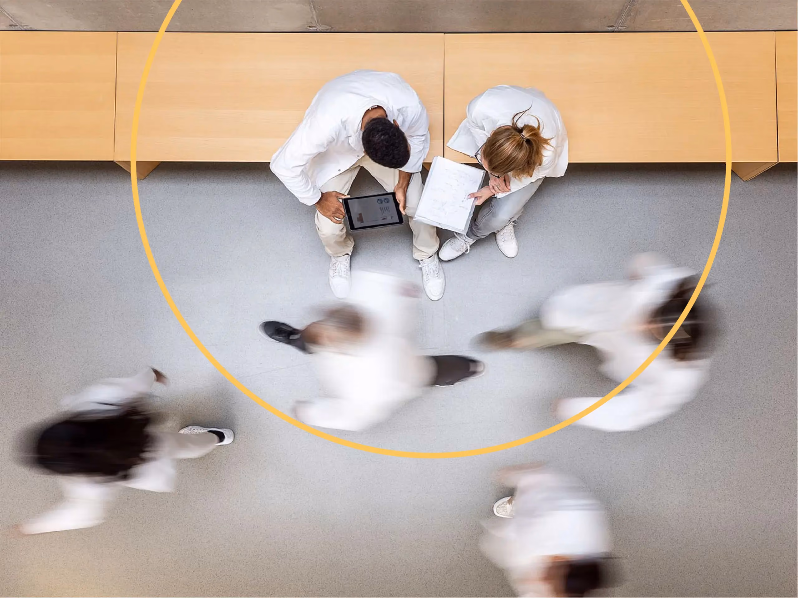 Overhead view of two people in white coats reviewing documents while others walk past in motion blur, framed by a yellow circle.