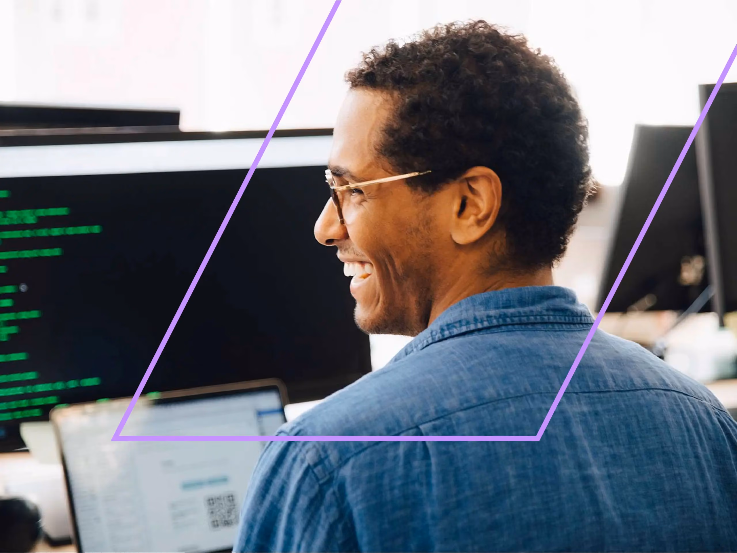 Man wearing glasses and a denim shirt smiling while looking at a computer monitor displaying code.