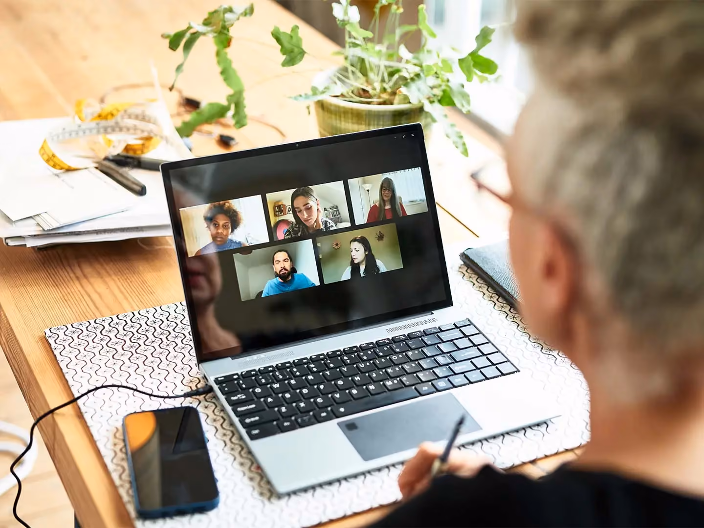 Person with gray hair participates in a video call with five people on a laptop screen, at a desk with plants and papers.