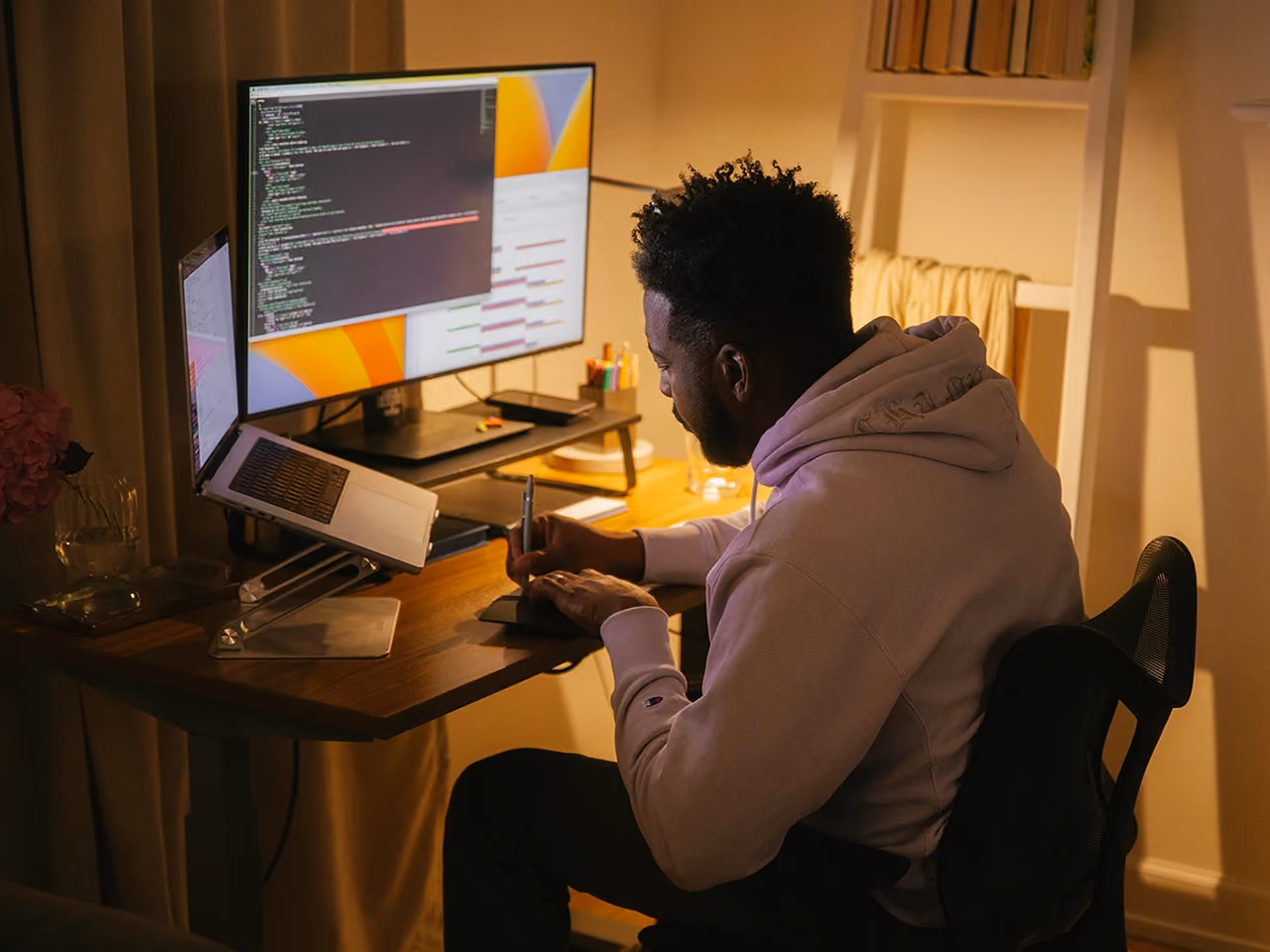 Man in a hoodie working at a desk with a laptop and monitor displaying code, in a dimly lit room.