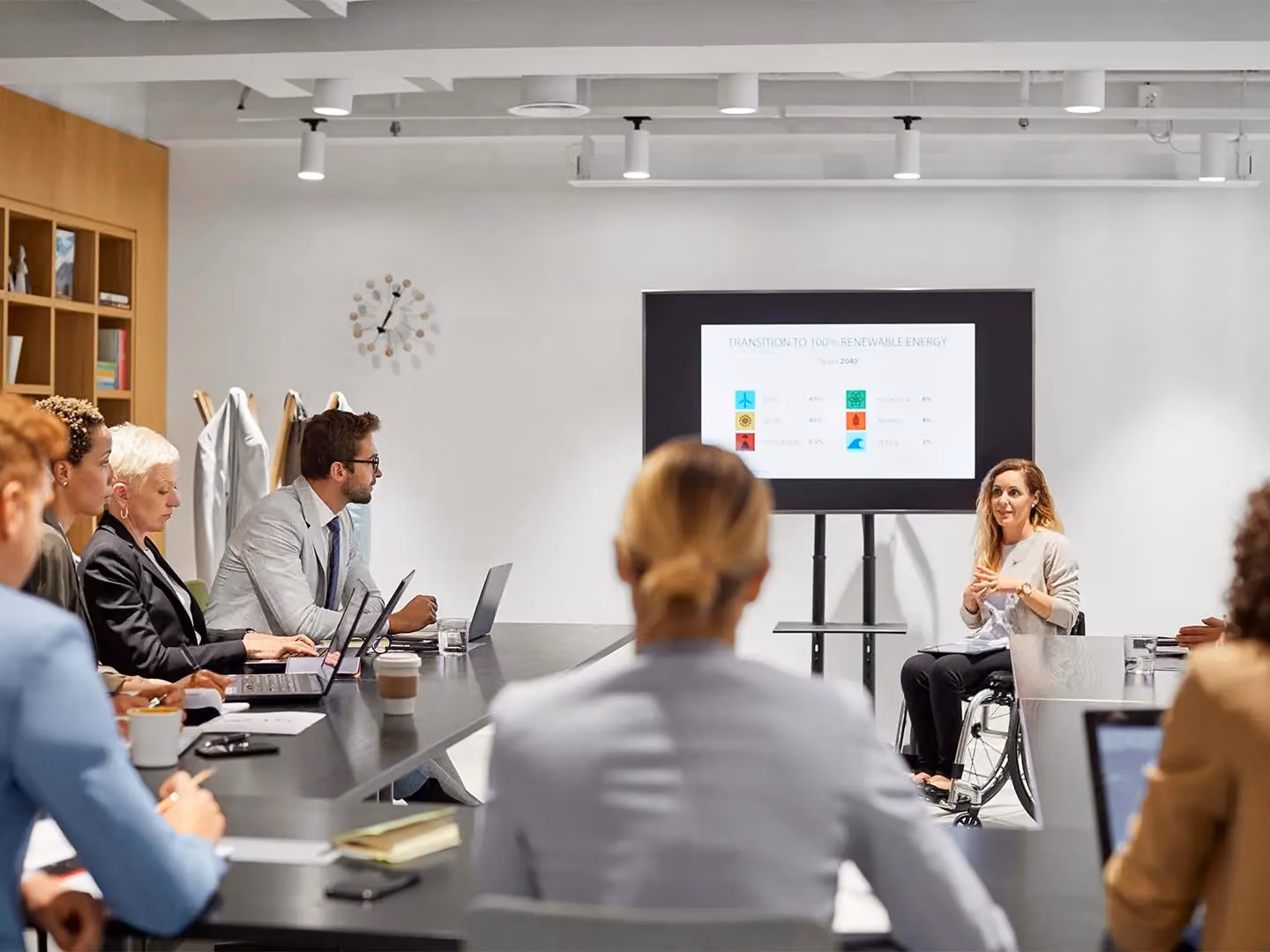 Woman using a wheelchair presents "Transition to 100% Renewable Energy" slide to colleagues in a conference room.