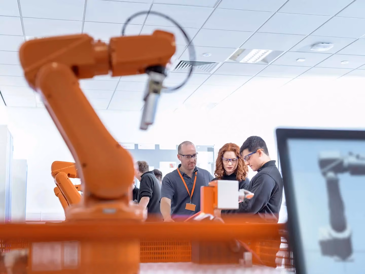 Three engineers wearing safety glasses examine equipment near an orange industrial robotic arm in a lab setting.