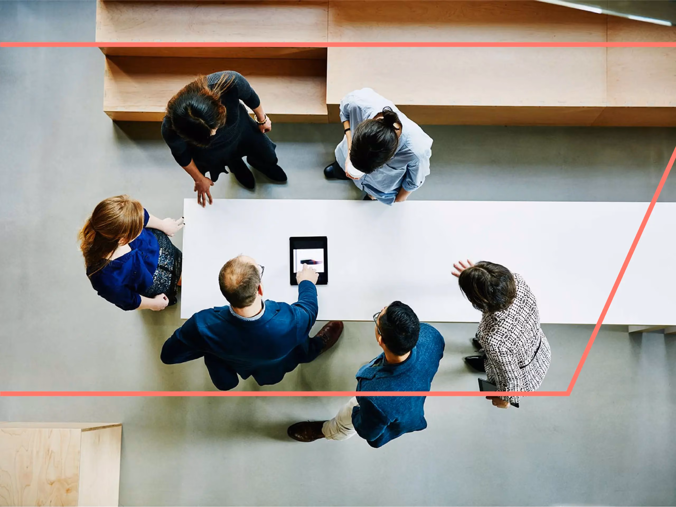 Overhead view of six people standing around a white table, gathered around a tablet device.