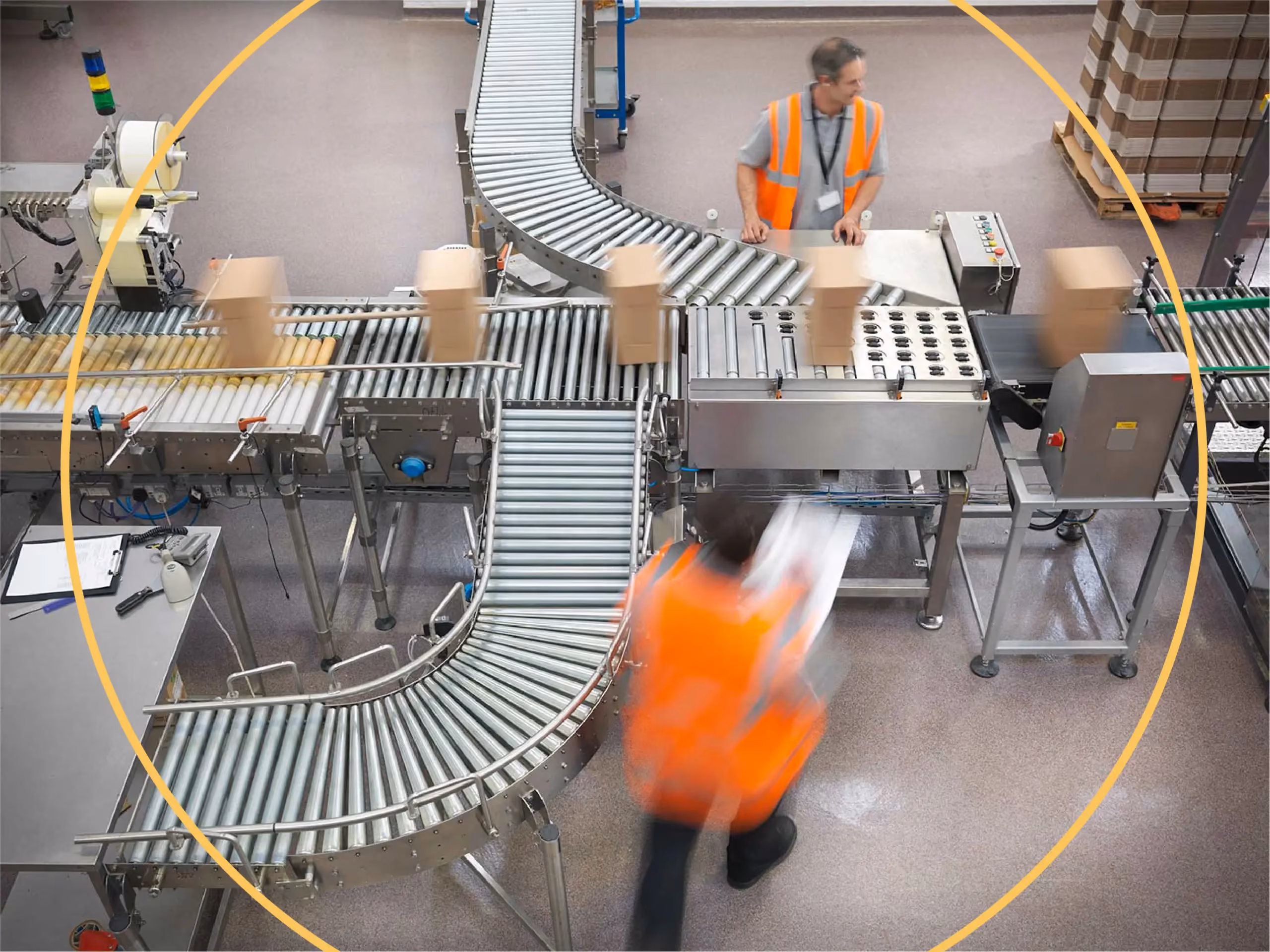 Overhead view of a warehouse conveyor belt system moving cardboard boxes, with two workers in orange safety vests nearby.