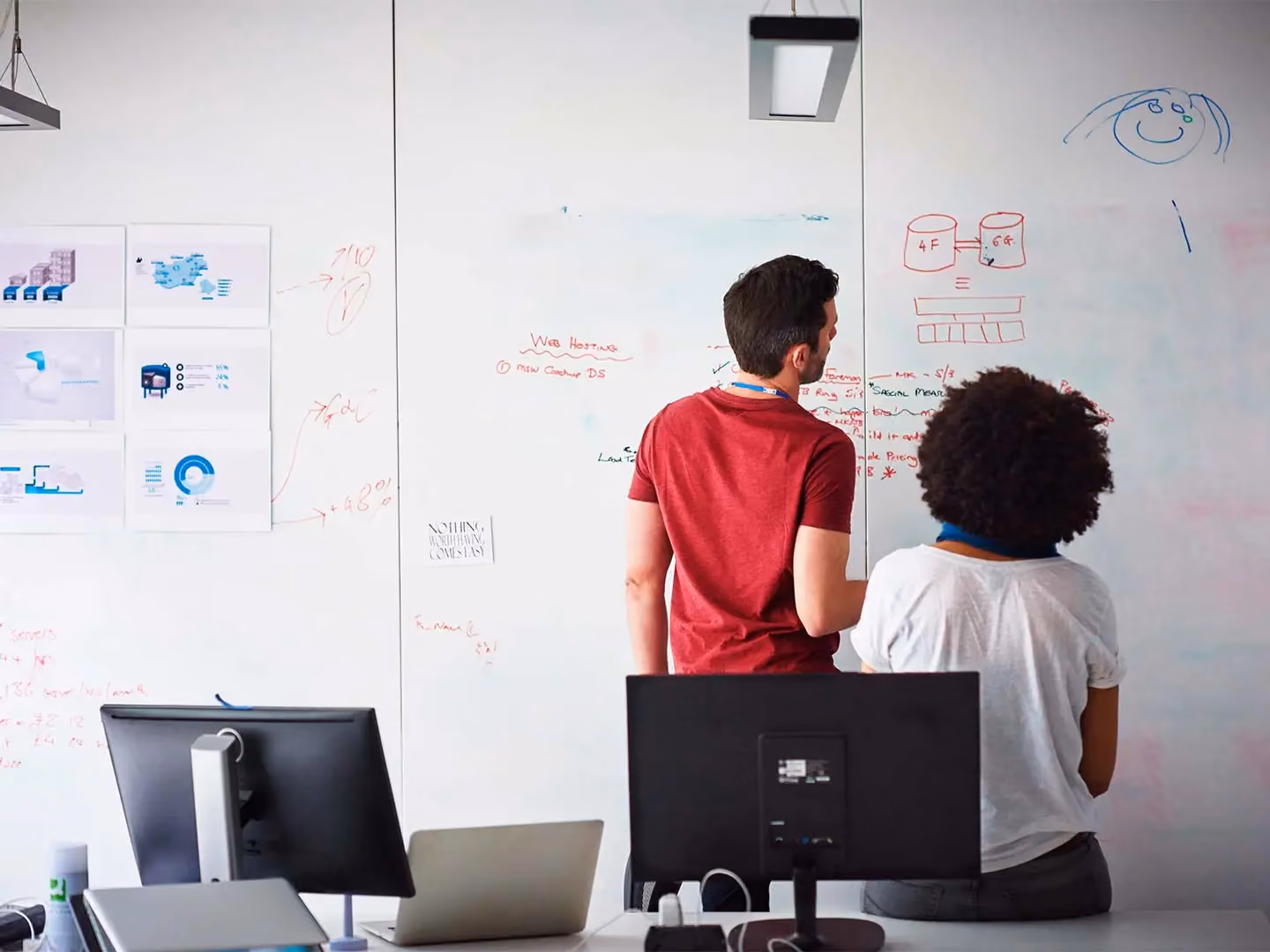 Two colleagues stand facing a whiteboard covered in diagrams and notes, including "Web Hosting" and printed charts on the left wall.