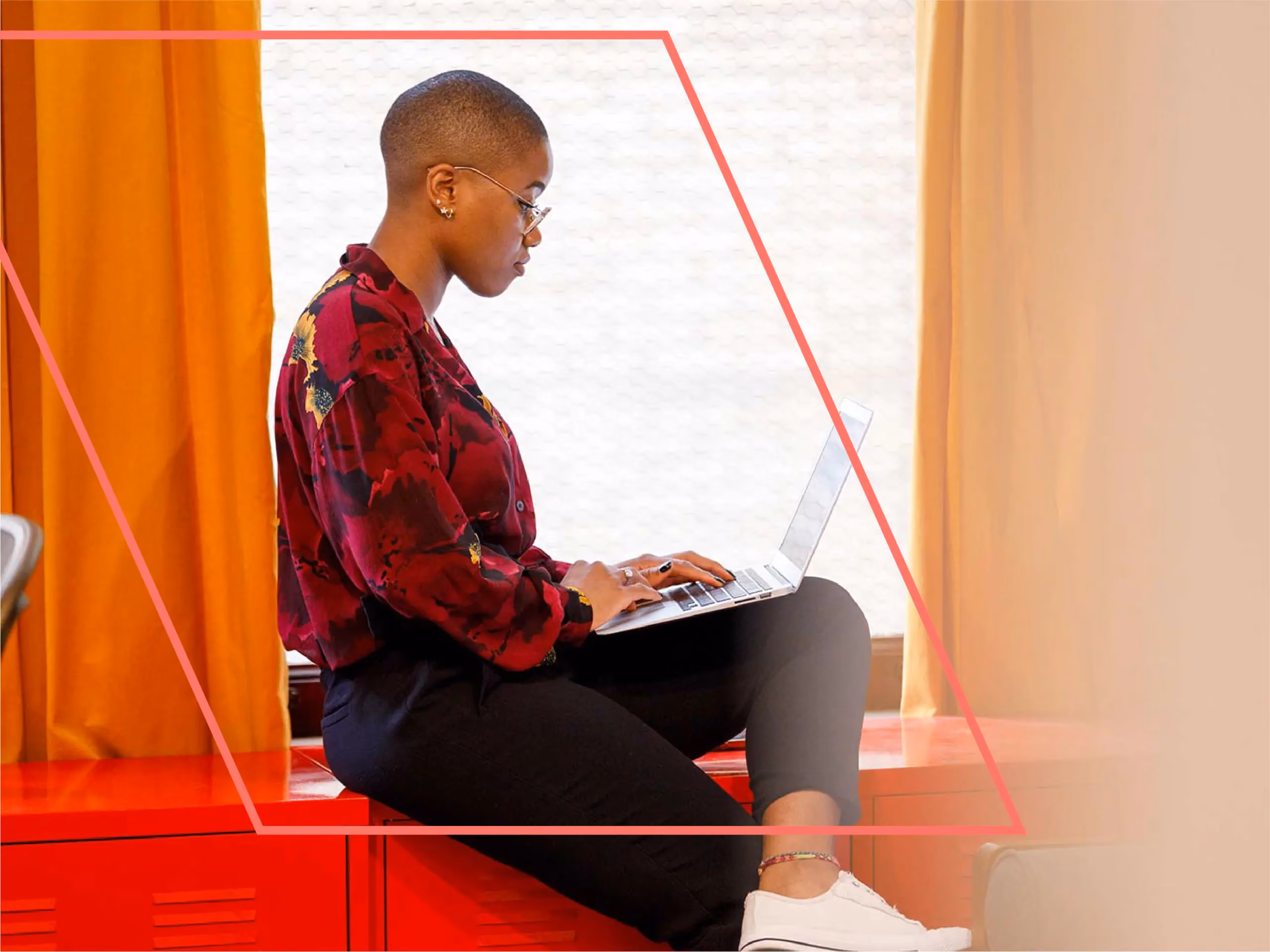 Woman with glasses sits on a red cabinet by a window, typing on a laptop.