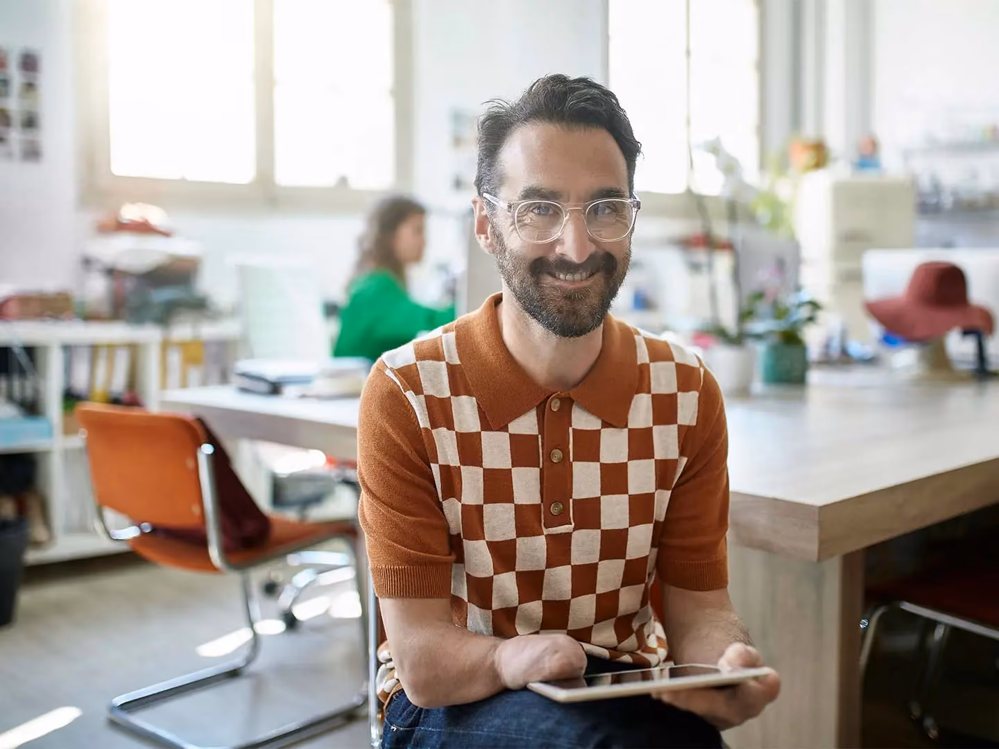 Man in orange checkered polo shirt holding a tablet, smiling in an office workspace with a colleague in the background.