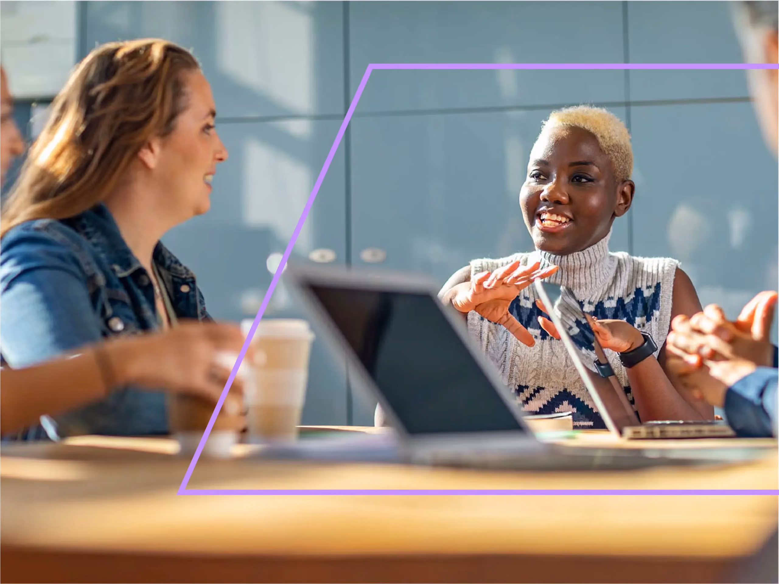Two women having an animated conversation at a table with laptops and coffee cups during a meeting.
