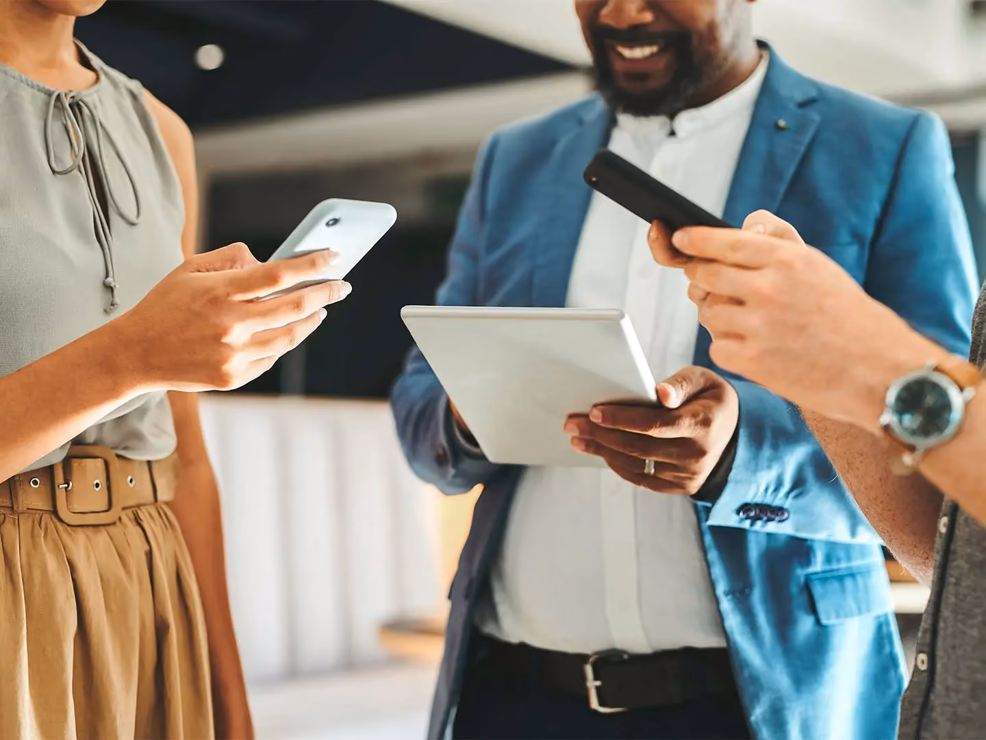 Three colleagues stand together, one holding a tablet, and two others holding smartphones.
