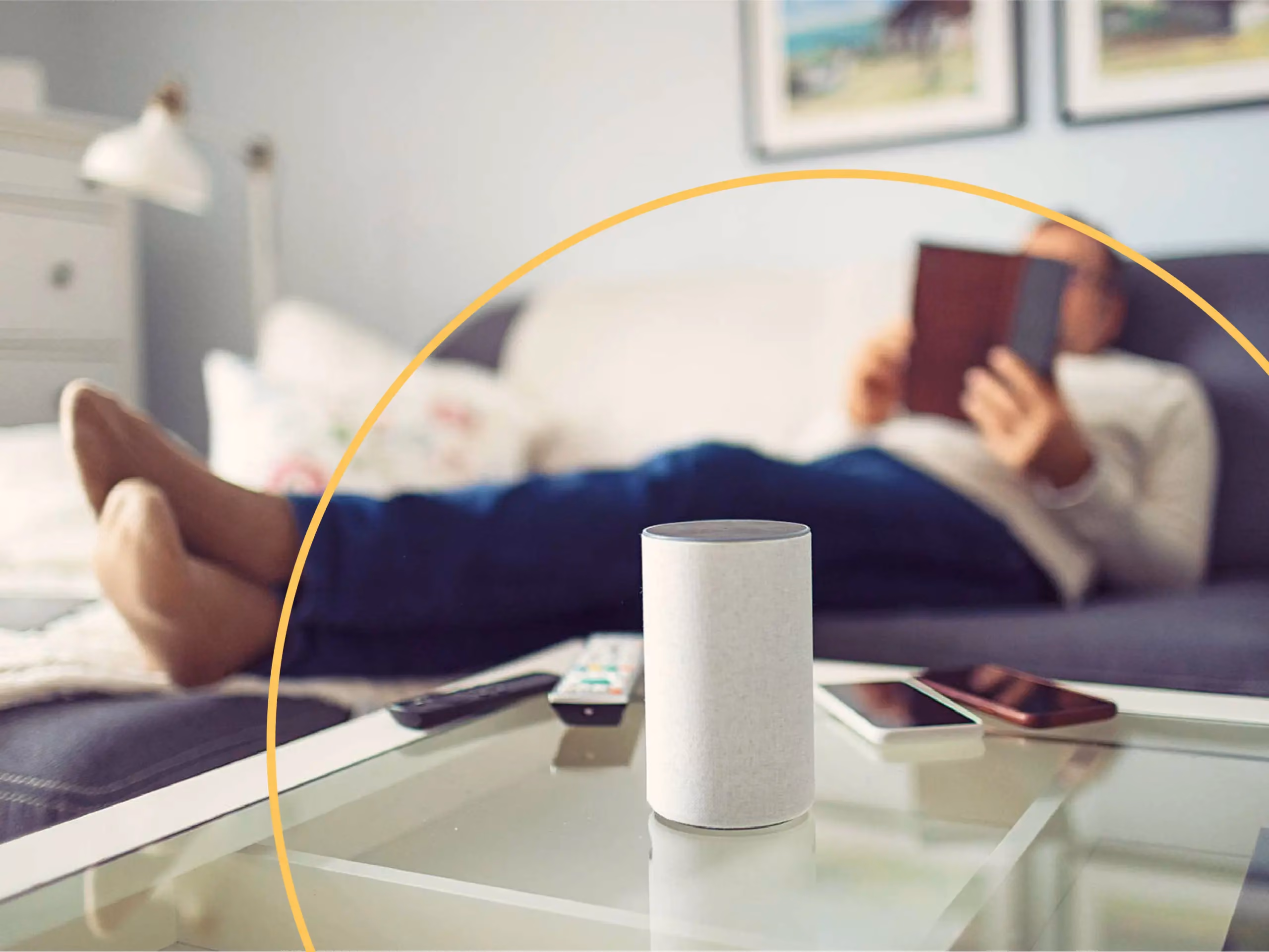 Smart speaker on a glass coffee table with remotes and phones, man reading a book on a couch in the background.