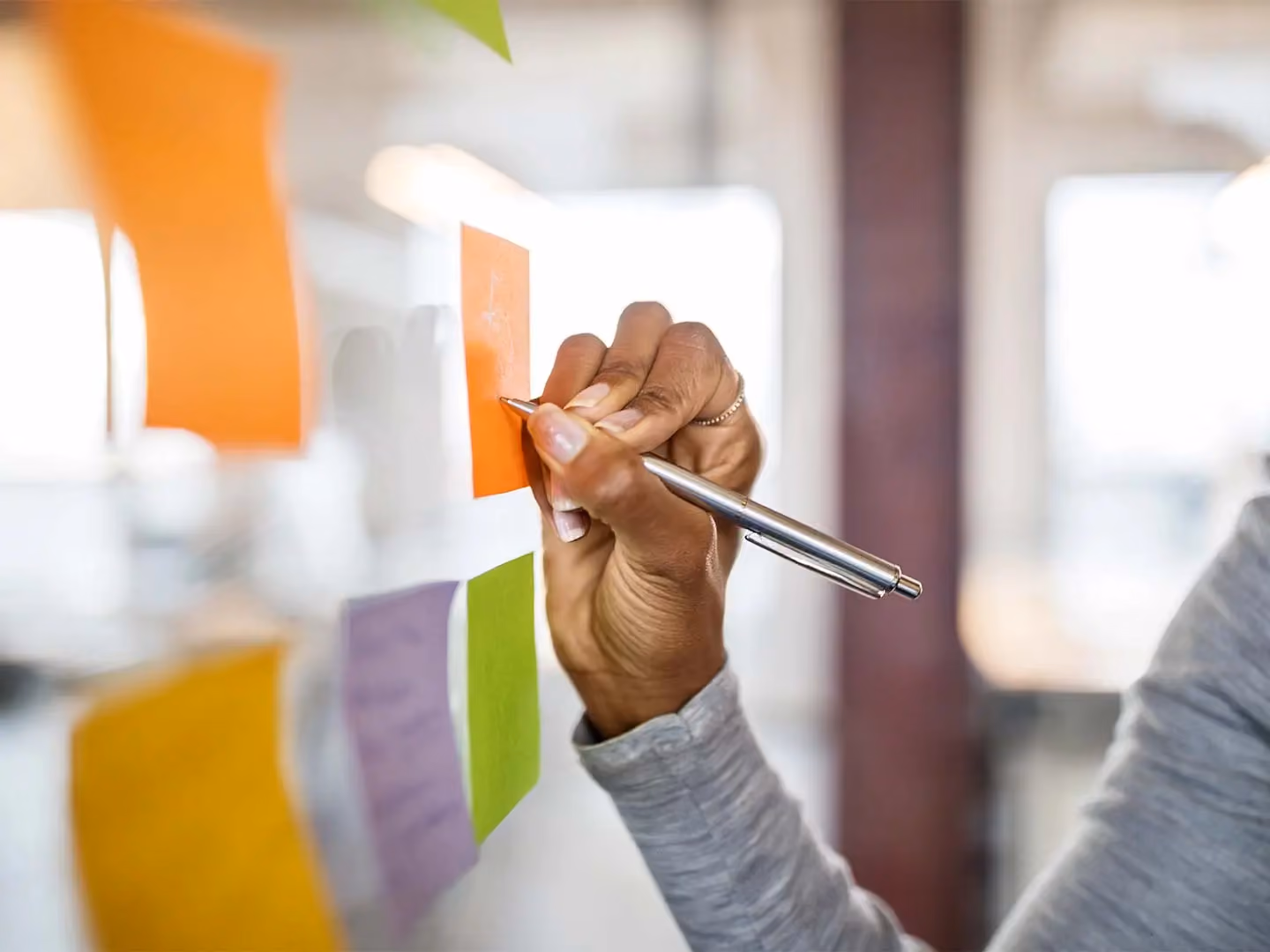 A hand holds a pen while writing on an orange sticky note among colorful notes on a glass surface.