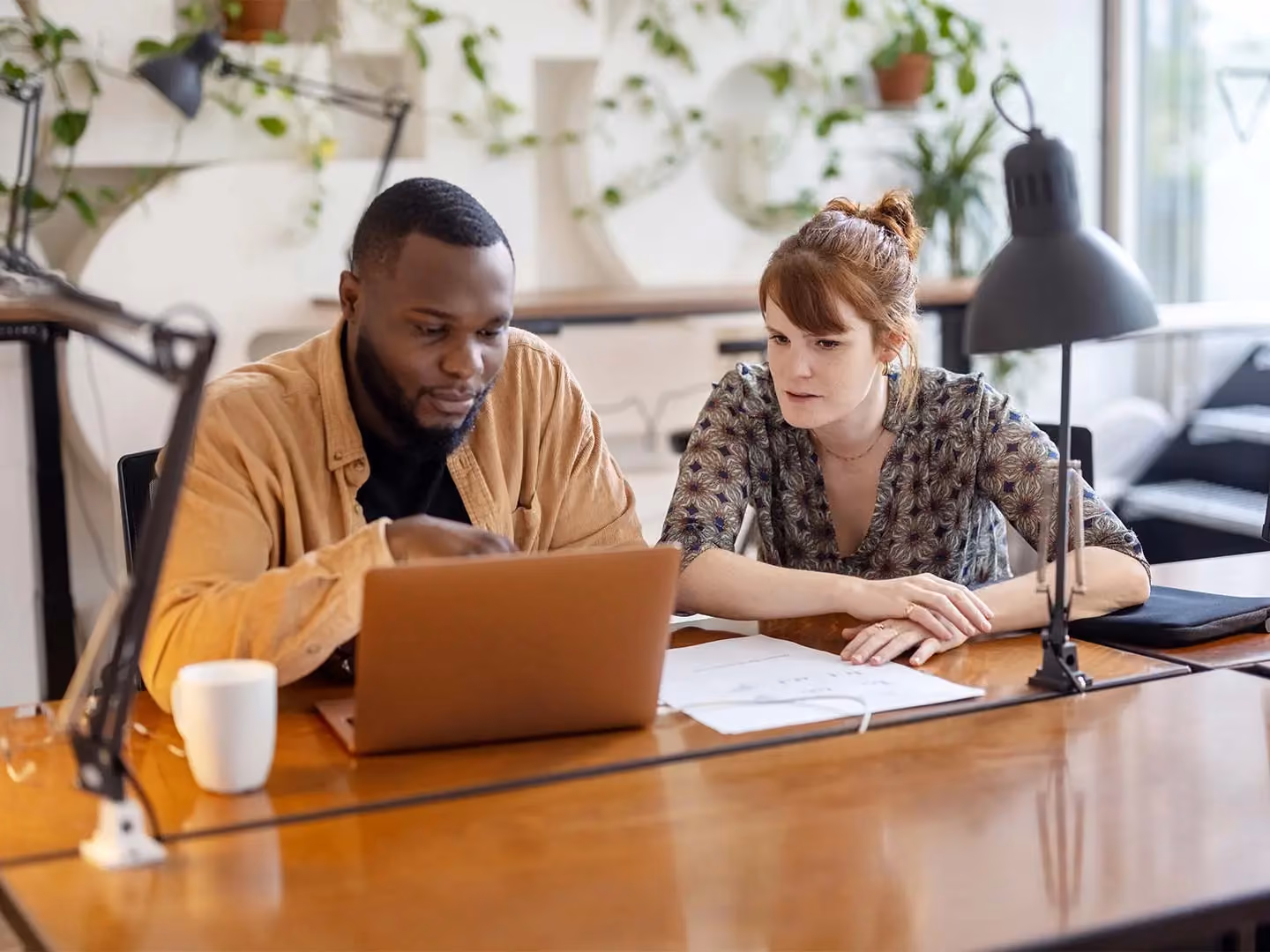 Man and woman sitting at a wooden desk looking at a laptop together, with papers, a mug, and desk lamps nearby.