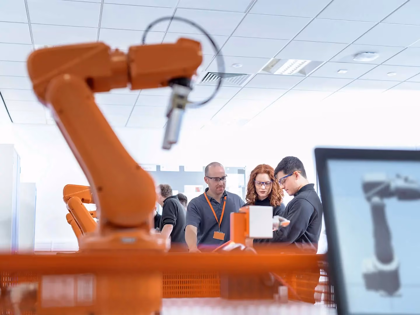 Three people wearing safety glasses examine equipment near an orange industrial robotic arm, with a monitor displaying its schematic.