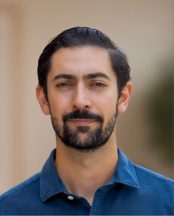 Man with dark hair and beard wearing a blue collared shirt, smiling slightly against a neutral background.