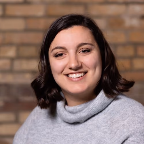 Woman with shoulder-length dark hair, smiling, wearing a gray turtleneck sweater, in front of a brick wall.