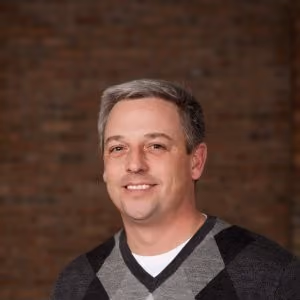 Headshot of a smiling man with short gray hair wearing a black and gray argyle sweater over a white shirt.