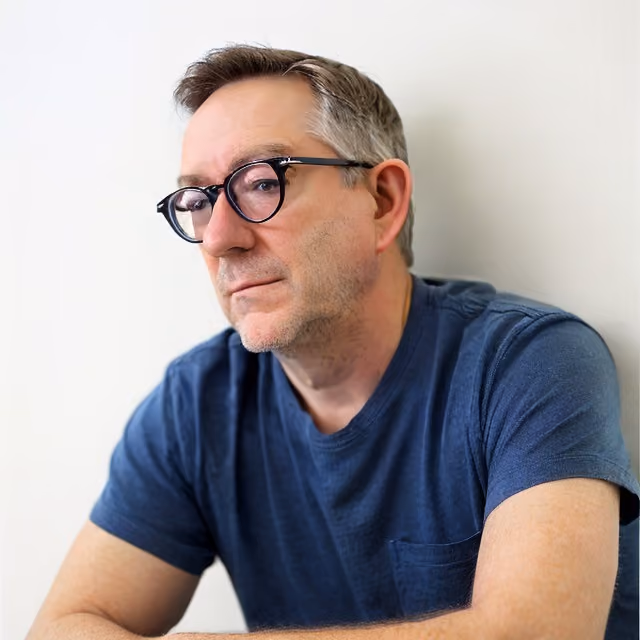 Man with short gray hair and dark-framed glasses wearing a navy blue T-shirt, seated against a light wall.