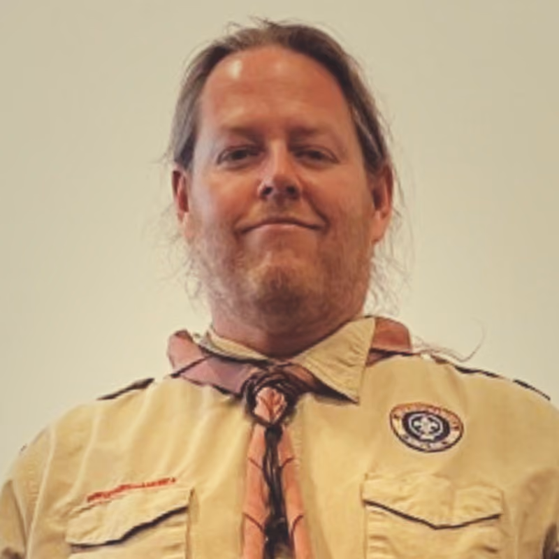 Man with a beard and ponytail wearing a tan scouting uniform with a neckerchief and emblem patch.