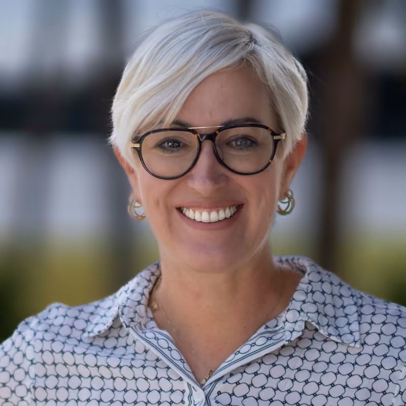 Woman with short blonde hair and glasses smiling in a patterned collared shirt, outdoor blurred background.