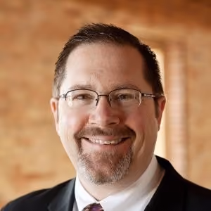 Headshot of a man with glasses and a beard, wearing a dark suit and patterned tie, in front of a brick wall.