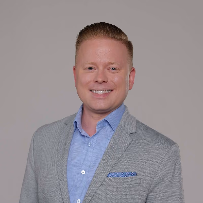 Professional headshot of a smiling man in a gray blazer and light blue shirt against a neutral background.