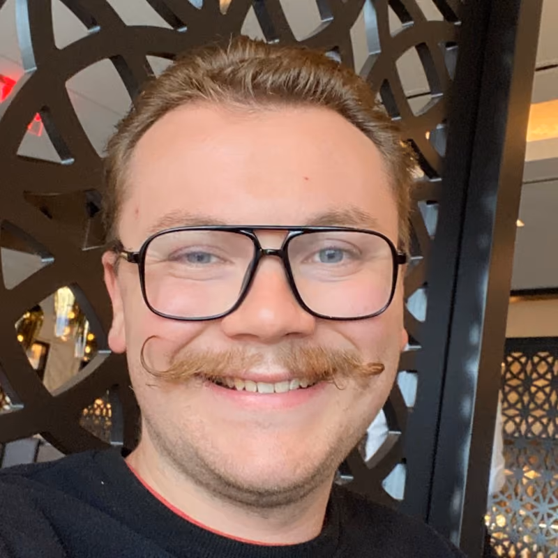 Man with a curled mustache and black-framed glasses smiling, wearing a black top, in front of a decorative screen.