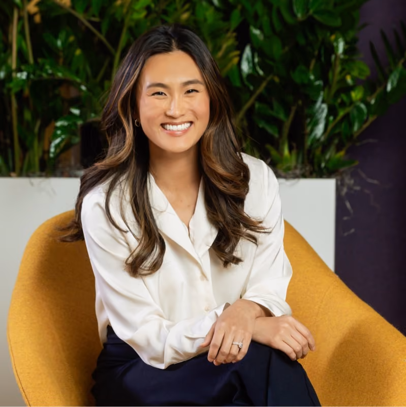 Woman with long brown hair wearing a white blouse sits smiling in a mustard yellow chair, plants in background.