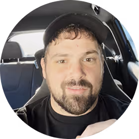 Man with a beard and mustache wearing a black cap and shirt inside a car.