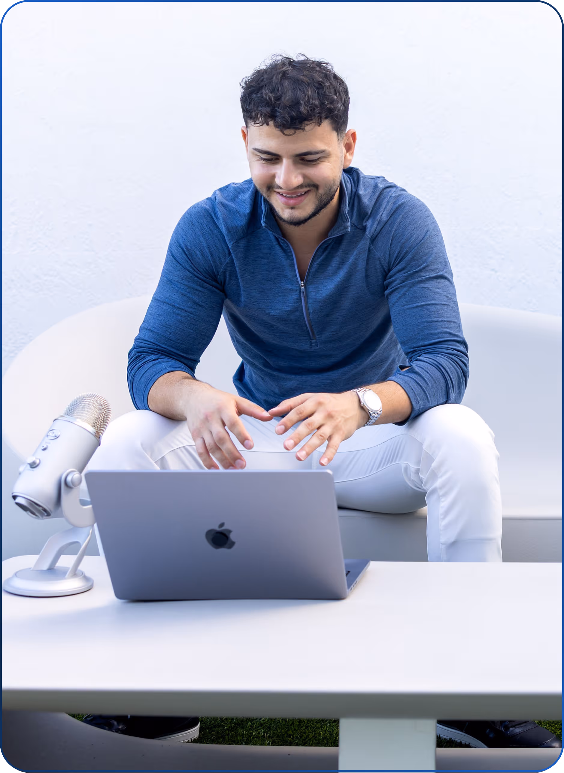 Young man in blue shirt sitting on couch, smiling at laptop with microphone on table.