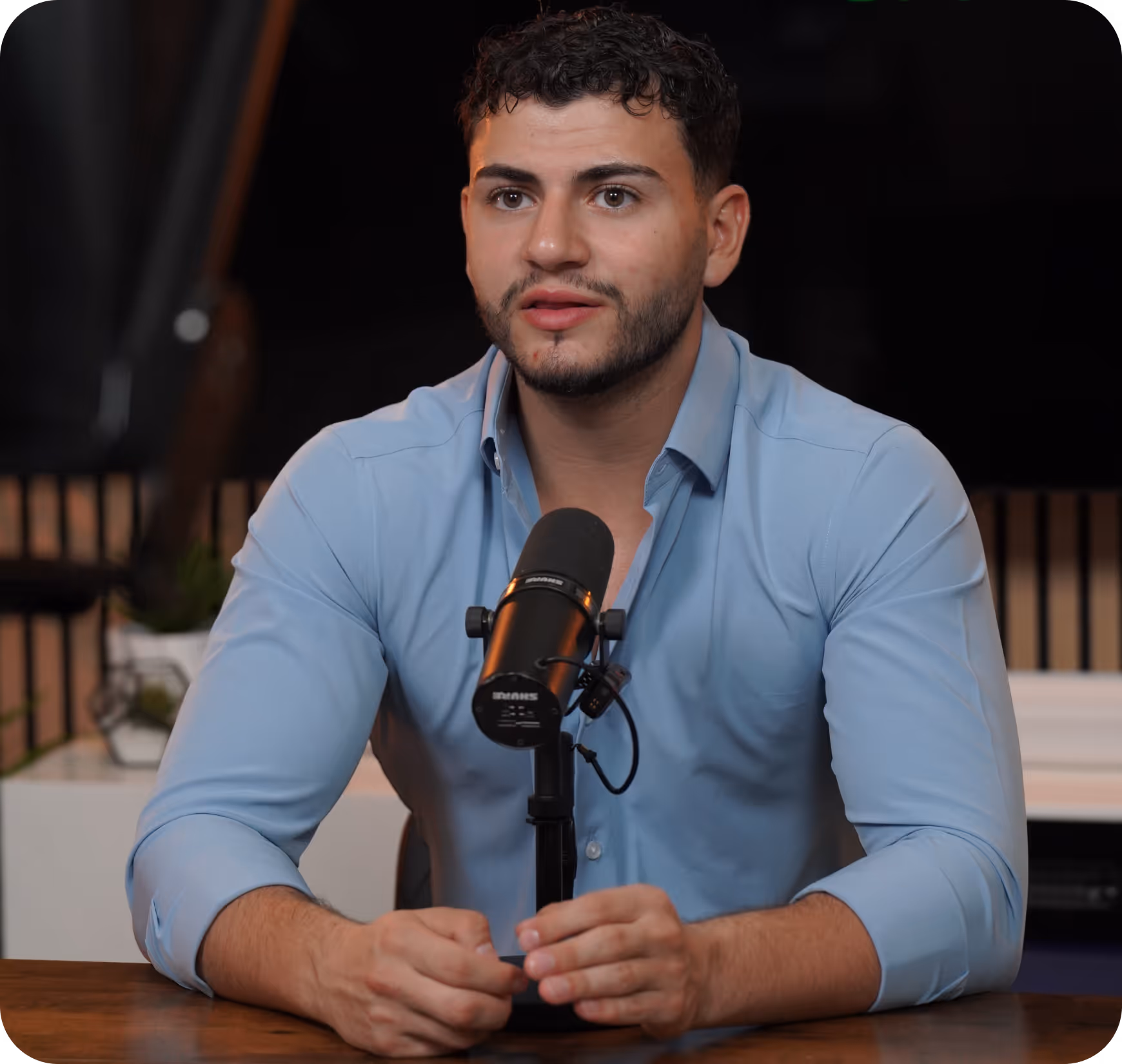 Man in blue shirt speaking into a tabletop microphone with hands clasped on a wooden table.