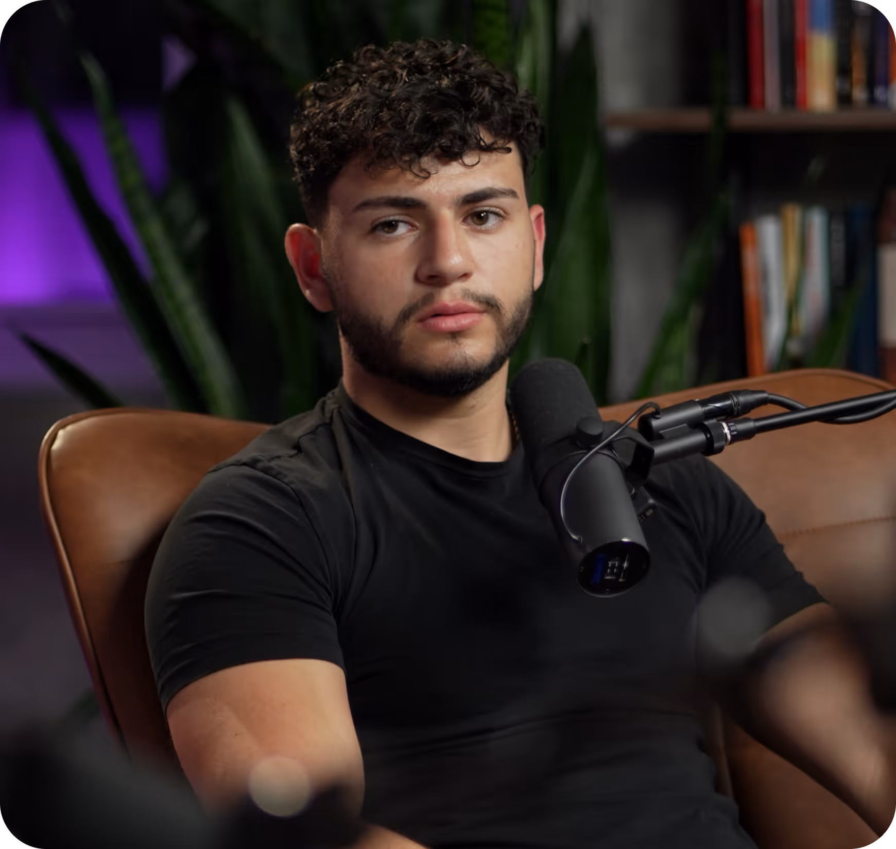 Young man with curly hair and beard speaking into a microphone, seated in a chair indoors.
