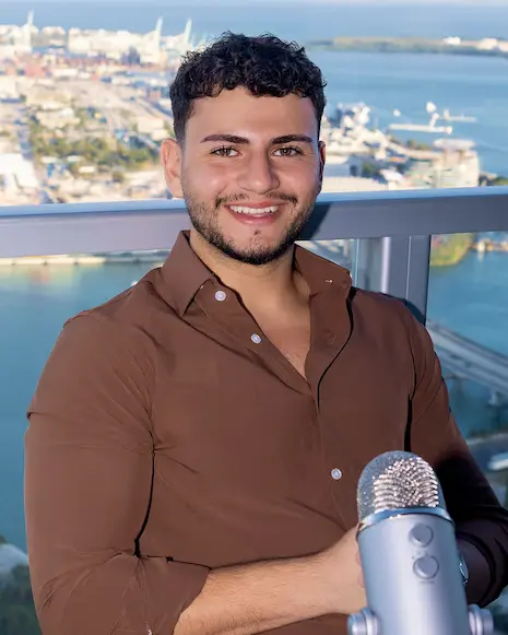 Smiling man in brown shirt in front of a microphone with a waterfront city view behind him.