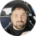 Smiling man with dark hair and beard wearing headphones inside a car.