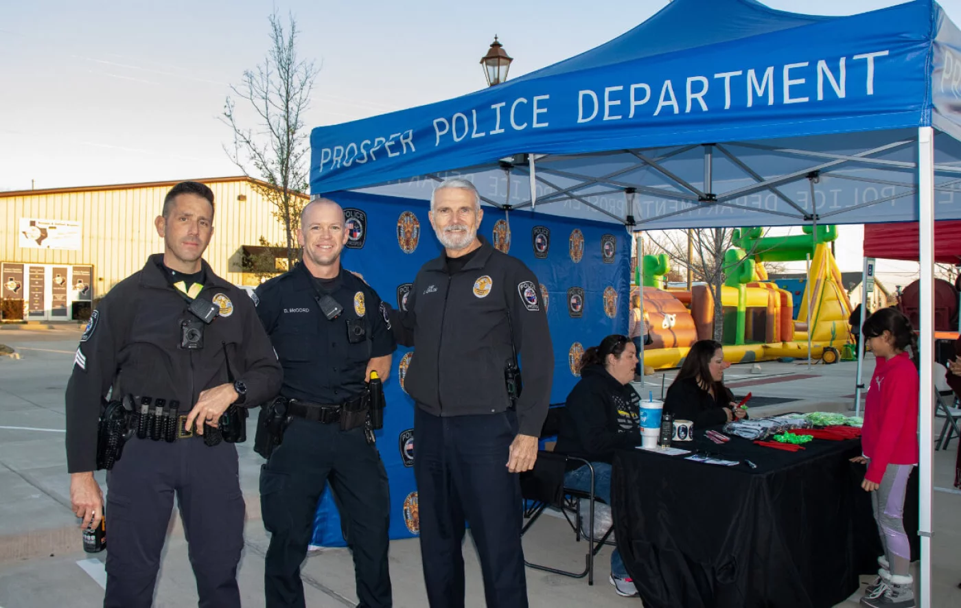 Three police officers standing in front of a Prosper Police Department tent with people at a table nearby.