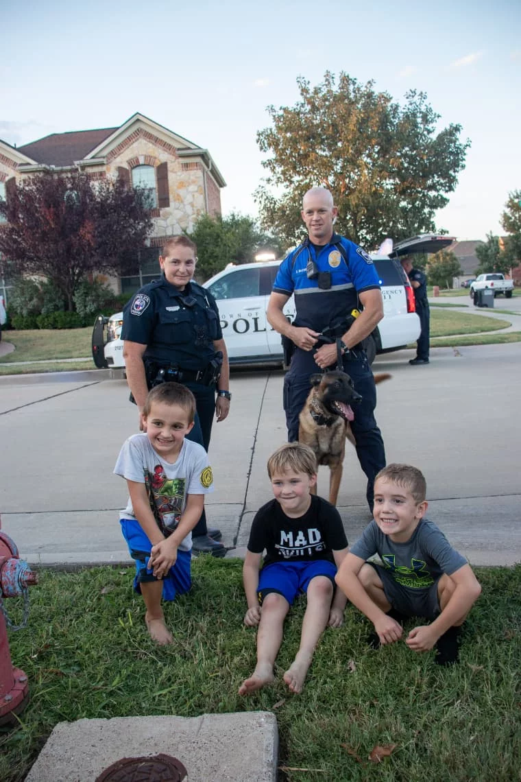 Two police officers and a K9 dog with three smiling boys sitting on grass in a neighborhood.