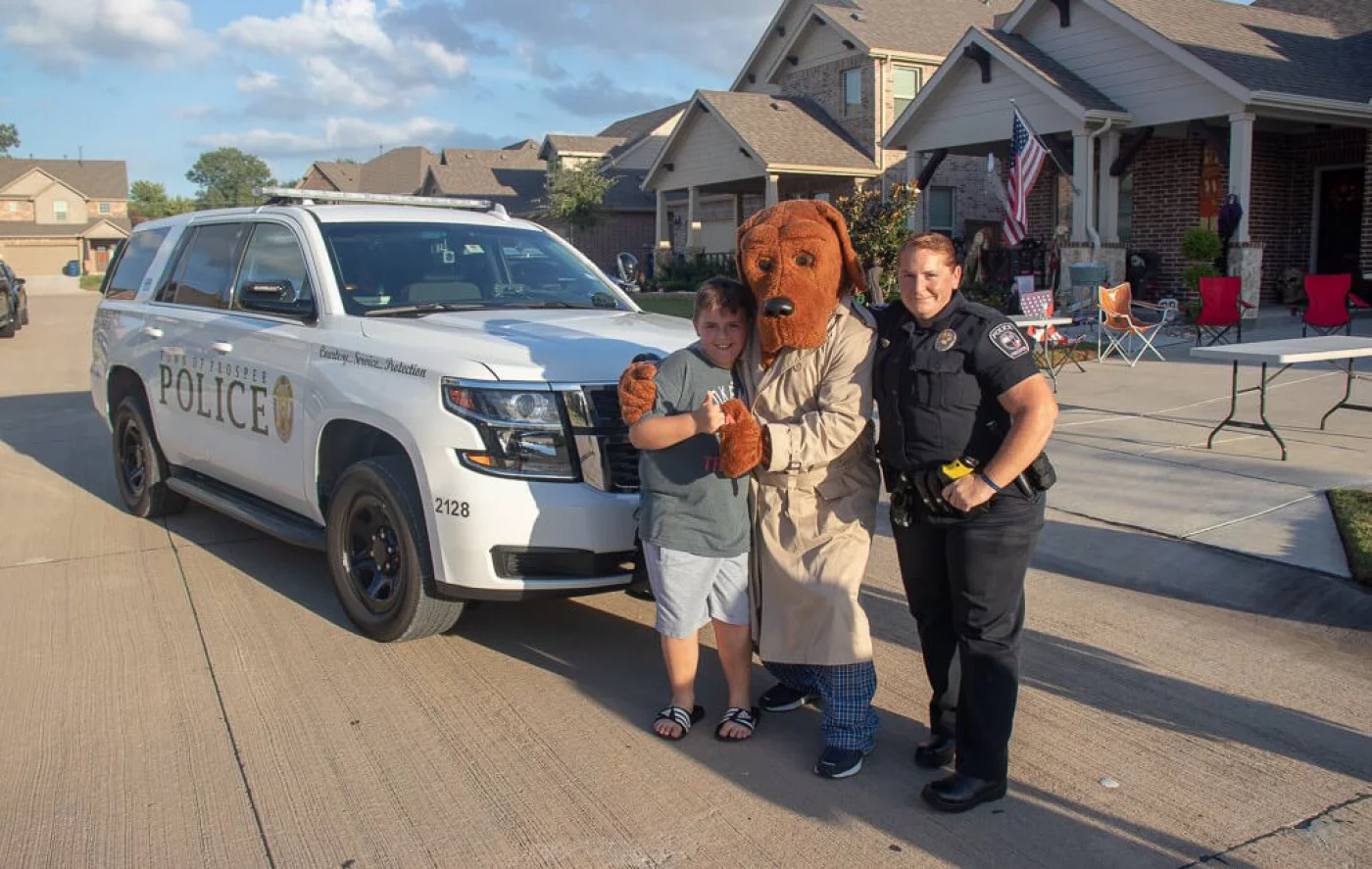 Child, person in dog mascot costume, and police officer posing next to a white police SUV on a suburban street.